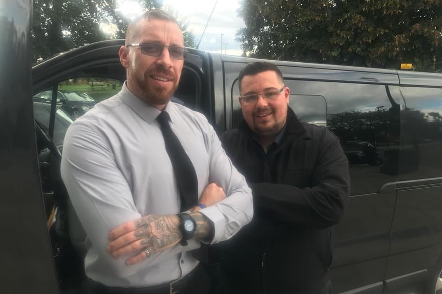 2 men dressed smartly with arms folded, standing in front of a black van