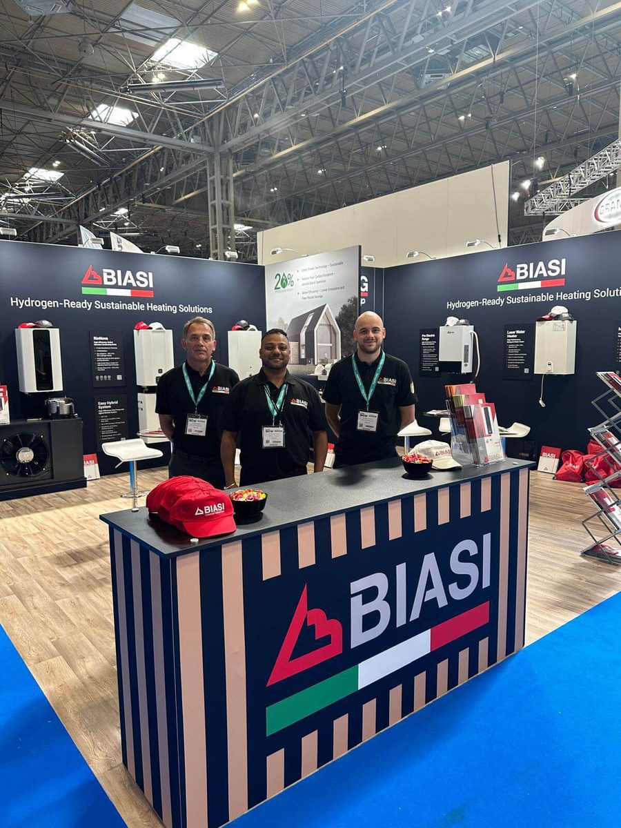 Three Biasi workers stood inside of the Biasi stand at the Installer Show. The stand has dark walls and the Biasi logo. Biasi boilers are mounted to the walls.