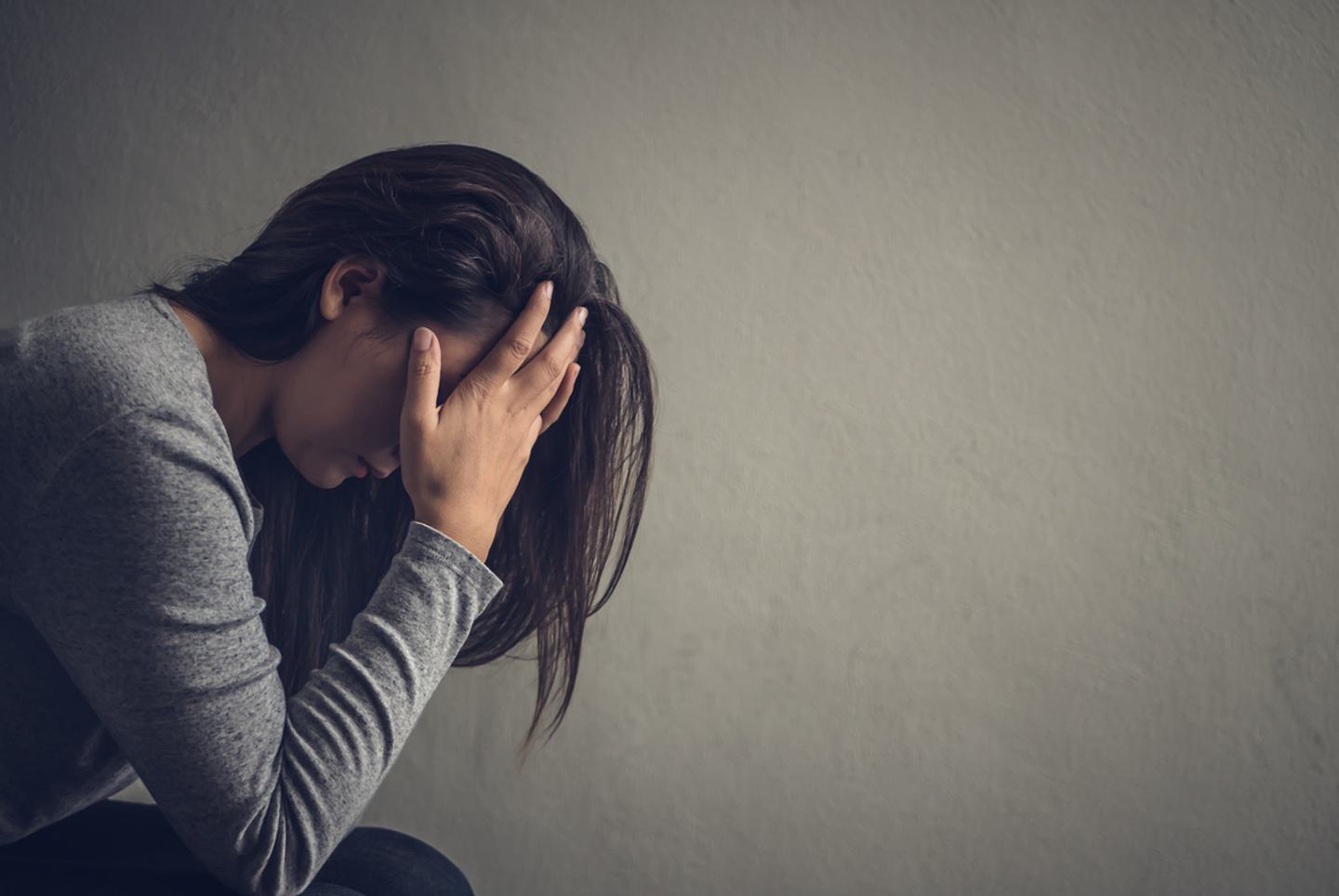 Woman sitting down, with her head in her hands. She is wearing a grey jumper and the background is grey creating a sombre mood.