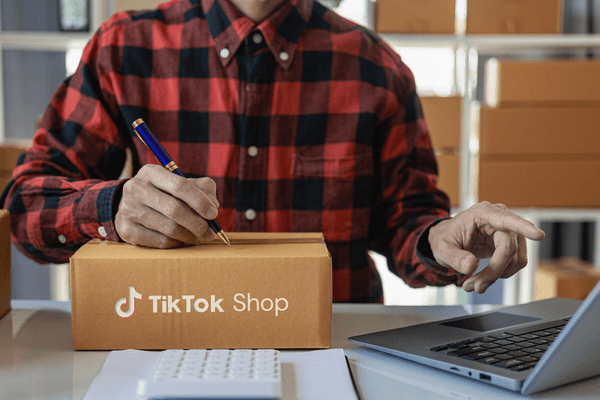 A man writing down shipping information on a carboard box for social selling, reading TikTok Shop. He is wearing a checked shirt standing behind the packing desk.