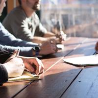 Team members having a discussion around a wooden table. Their faces are mostly out of frame, however you can see their hands and notepads. There is a glass window at the end of the table flooding light onto the table.