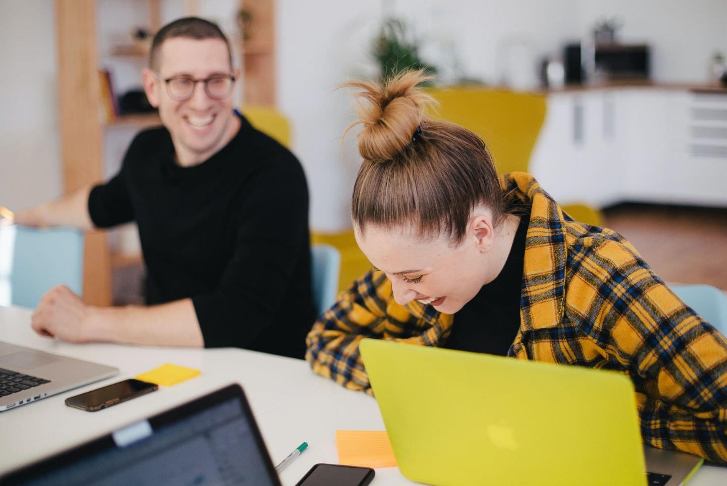 An image of 2 work colleagues laughing at something funny. The man is in a black jumper looking at his co-worker and laughing. His co-worker is female and she is wearing a yellow and black patterned shirt.
