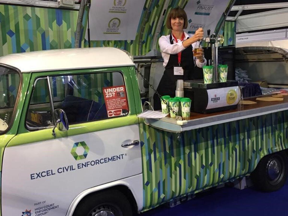 Classic VW camper van wrapped in Excel civil Enforcement’s branding. The roof is raised (pivoted from one side) and a lady is inside serving beer from a counter.