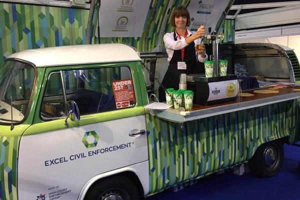 Classic VW camper van wrapped in Excel civil Enforcement’s branding. The roof is raised (pivoted from one side) and a lady is inside serving beer from a counter.