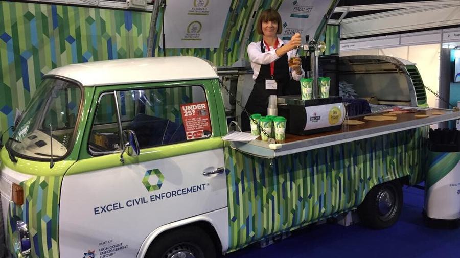 Classic VW camper van wrapped in Excel civil Enforcement’s branding. The roof is raised (pivoted from one side) and a lady is inside serving beer from a counter.