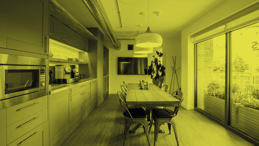 Interior view of a kitchen area. A long galley table sits centrally lit by multiple overhead handing lights. A kitchen covers the entire length of the room (on the left) with full height windows on the right. The image has a yellow filter applied.