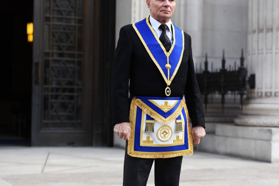 A Freemason standing in black suit and tie wearing a white shirt and wearing the Freemason apron and sash. His sash is a royal blue with gold trim as is the apron that sits on his waist with golden tassels and a circular logo with London embroidered on.