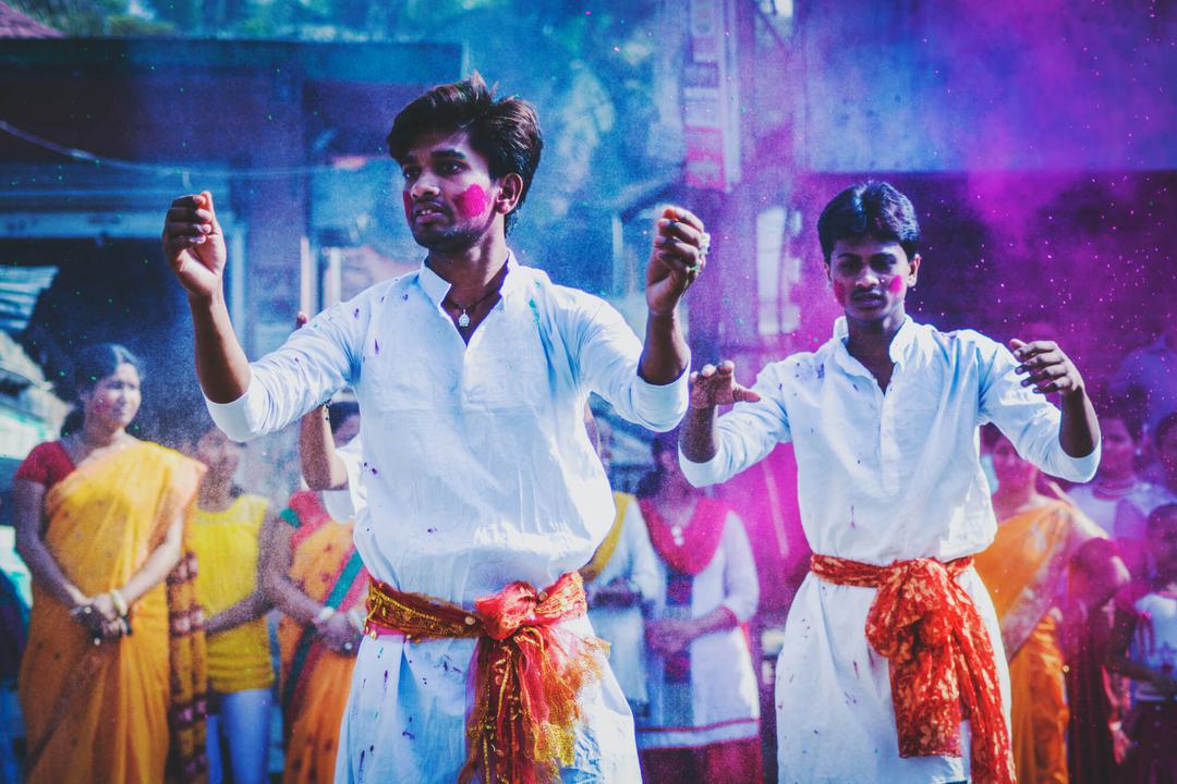 A colourful image of men dancing an wearing face paint. There are vibrant colours all around