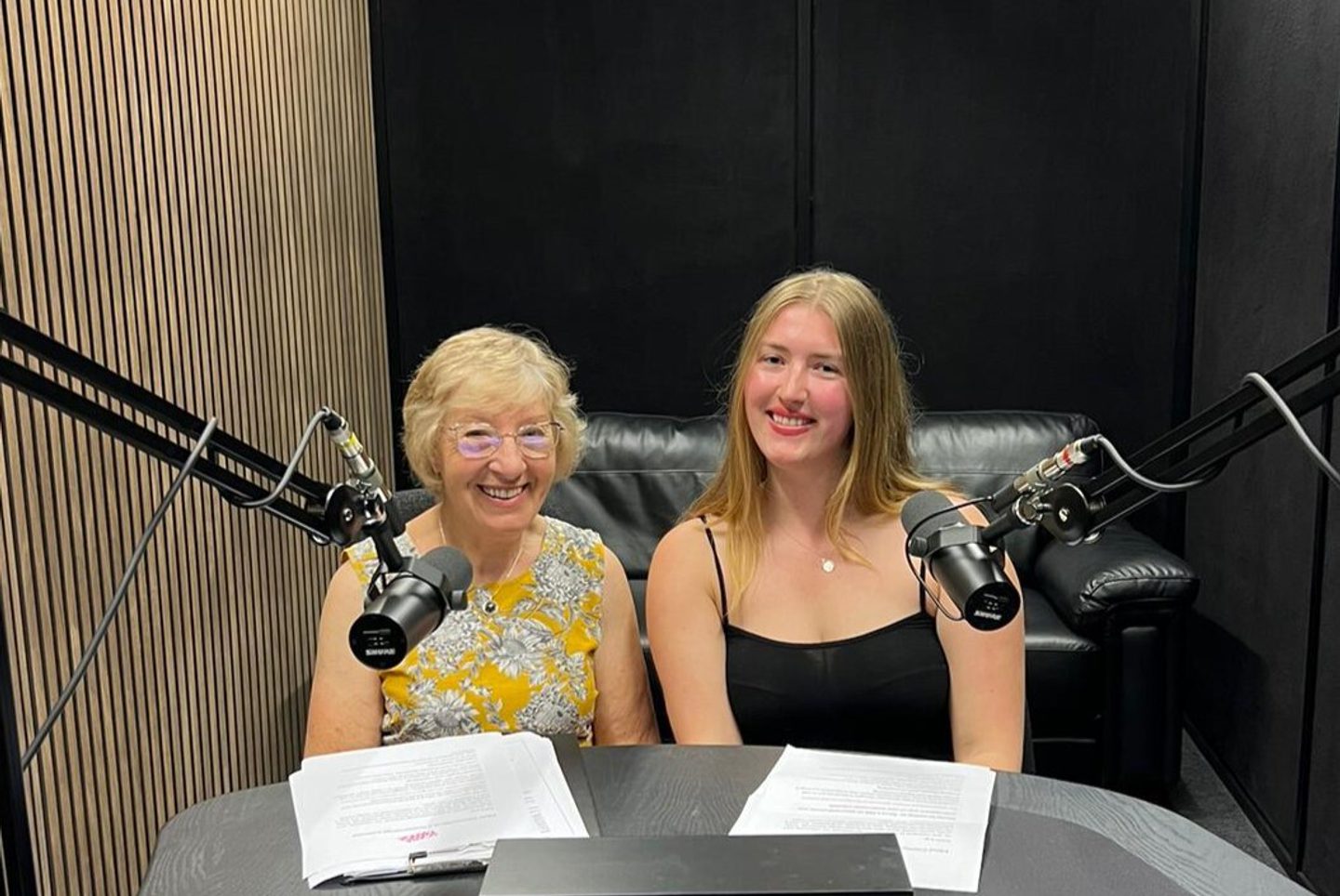 Pat and Charlotte sat in the podcast recording studio. Pat is 82, she has light hair and is wearing a flowery dress. Charlotte is in her mid-twenties, has long brown hair and is wearing a black dress.