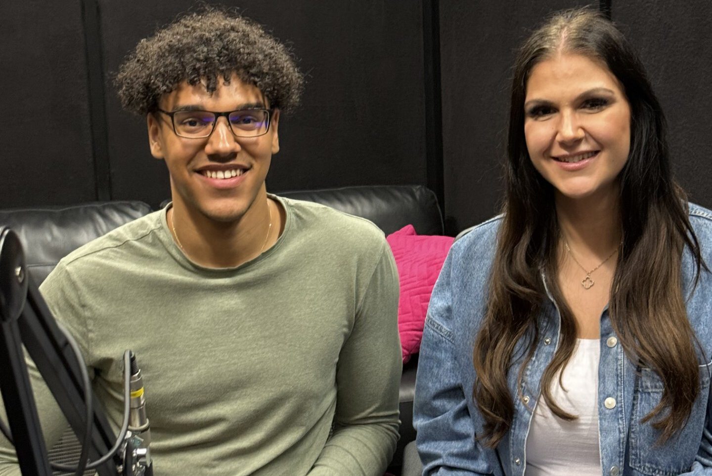 Jamie (left) and Emma (right) sitting at the desk in the podcast studio smiling; there is an ON AIR sign on the desk table